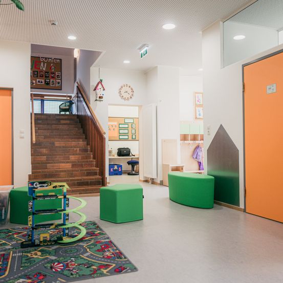 Ein Spielzimmer mit grünen Möbeln, einem Teppich und einer Treppe. Eine Uhr und ein Vogelhaus befinden sich an der Wand.