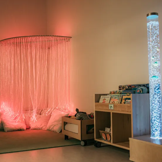 Eine gemütliche Leseecke mit einer gebogenen LED-Leuchte, einem Bücherregal und Spielzeug auf einem Teppich. Die Lichter schaffen eine warme Atmosphäre.