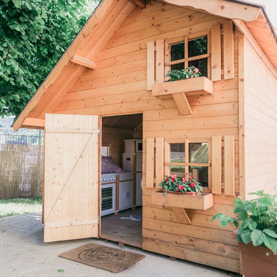 Ein hölzernes Spielhaus mit offener Tür, die eine kleine Küche im Inneren zeigt. Blumentöpfe stehen vor den Fenstern.