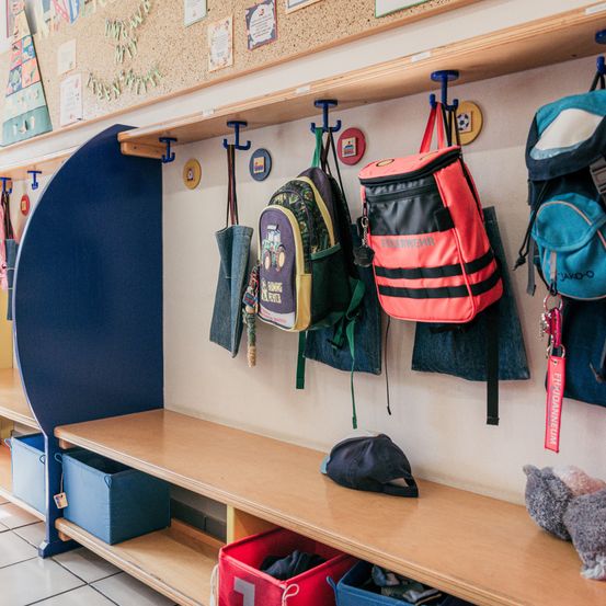 Ein Klassenzimmer mit verschiedenen Gegenständen an der Wand, darunter Rucksäcke, Hüte und Aufbewahrungsboxen, mit einem Holzregal und einer blauen Aufbewahrungsbox.