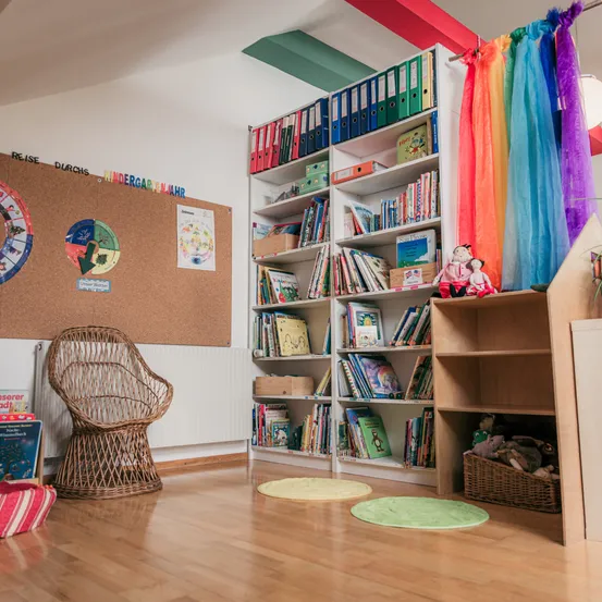 Ein gemütliches Zimmer mit Holzboden, einem Korbstuhl und einer Pinnwand. Ein Bücherregal mit bunten Büchern und Ordnern steht neben einem Regal mit Spielzeug. Bunte Schals hängen von der Decke.