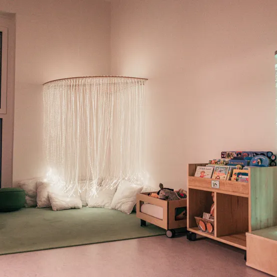 Ein gemütliches Spielzimmer mit rosa Wänden, einem weißen Vorhang, Kissen, Spielzeug und einem Holzregal voller Bücher.