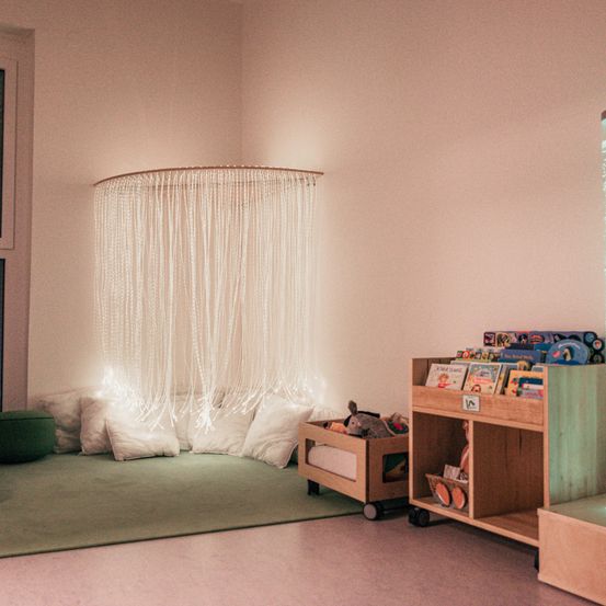 Ein gemütliches Spielzimmer mit rosa Wänden, einem weißen Vorhang, Kissen, Spielzeug und einem Holzregal voller Bücher.