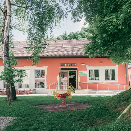 Ein orangefarbenes Gebäude mit einem braunen Ziegeldach, umgeben von Bäumen, hat ein Schild mit der Aufschrift 'KINDER'. Davor steht ein roter und gelber Federpferd, und ein Rasenbereich mit einer Rampe, die zum Eingang führt.