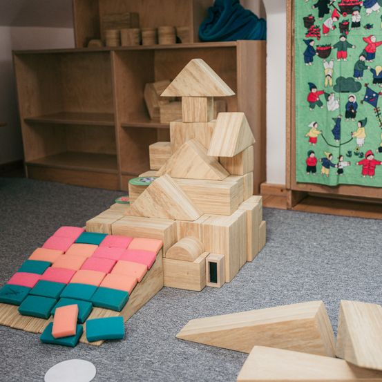 Ein Raum mit Holzregalen und einem Teppichboden zeigt eine große Holzblockstruktur. Die Struktur besteht aus einem Dach, Wänden und verschiedenen Blöcken. Daneben befindet sich ein Haufen kleinerer Blöcke.