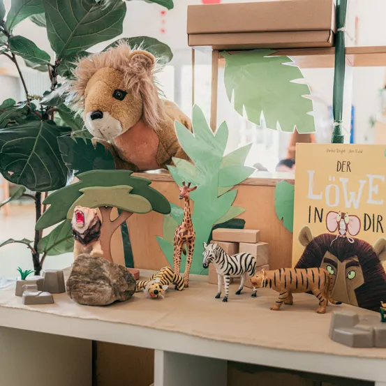 Ein Löwenplüschtier sitzt auf einem Regal. Verschiedene Spielzeugtiere und ein Buch mit dem Titel 'Der Löwe in Dir' sind ausgestellt. Die Umgebung ähnelt einem Dschungel mit Pappausschnitten und Felsen.