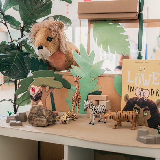 Ein Löwenplüschtier sitzt auf einem Regal. Verschiedene Spielzeugtiere und ein Buch mit dem Titel 'Der Löwe in Dir' sind ausgestellt. Die Umgebung ähnelt einem Dschungel mit Pappausschnitten und Felsen.