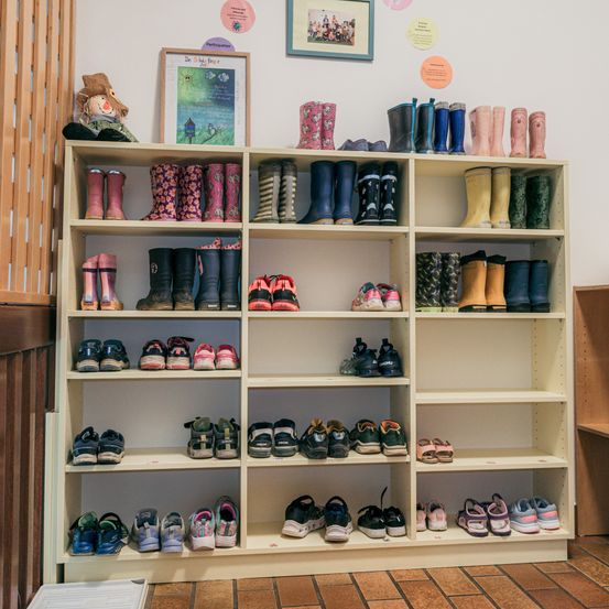 Ein Holzregal voller verschiedener Kinderstiefel und Schuhe, einschließlich Regenstiefel und Turnschuhe, an einer weißen Wand mit Bildern und Postern.