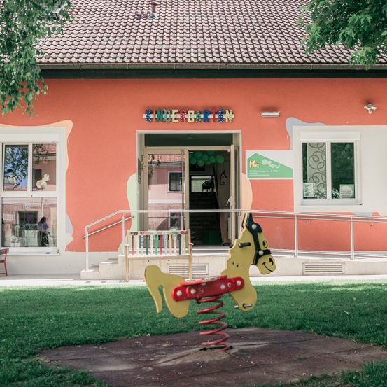 Die Vorderseite eines Kindergartens mit farbenfroher Außenseite, Glasfenstern und einem Schild mit der Aufschrift 'Kindergarten'. Vor dem Gebäude befindet sich ein Spielplatz mit einem bunten Pferdespielzeug.