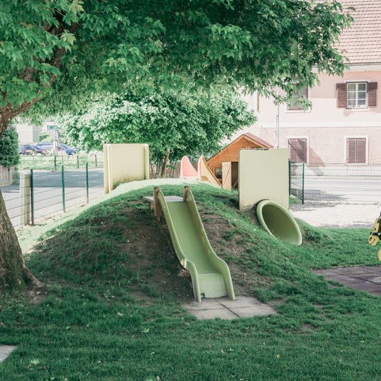 Ein Spielplatz mit Rutschen und einem kleinen Haus steht unter einem Baum mit grünen Blättern, umgeben von einem Zaun. Hinter dem Zaun befindet sich ein Gebäude mit Fenstern und einem geparkten Auto in der Nähe.
