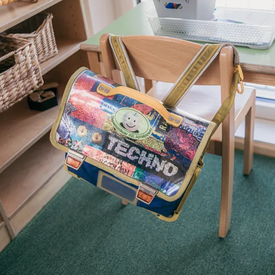Ein bunter Rucksack mit dem Wort 'Techno' darauf hängt an einem Holzstuhl. Er steht auf einem grünen Teppich. Dahinter befindet sich ein Holzschreibtisch mit einer Aufbewahrungsbox darauf. Im Hintergrund steht ein Holzregal mit Weidenkörben.