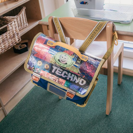 Ein bunter Rucksack mit dem Wort 'Techno' darauf hängt an einem Holzstuhl. Er steht auf einem grünen Teppich. Dahinter befindet sich ein Holzschreibtisch mit einer Aufbewahrungsbox darauf. Im Hintergrund steht ein Holzregal mit Weidenkörben.