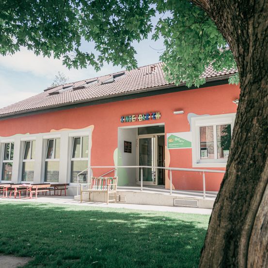 Ein rotes Gebäude mit weißen Verzierungen hat ein Schild mit der Aufschrift 'Kindergarten'. Es hat mehrere Fenster und eine Tür mit einem grünen Schild. Draußen gibt es eine Bank und Tische mit Stühlen. Das Gebäude ist von einer Rasenfläche umgeben.