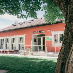 Ein rotes Gebäude mit weißen Verzierungen hat ein Schild mit der Aufschrift 'Kindergarten'. Es hat mehrere Fenster und eine Tür mit einem grünen Schild. Draußen gibt es eine Bank und Tische mit Stühlen. Das Gebäude ist von einer Rasenfläche umgeben.