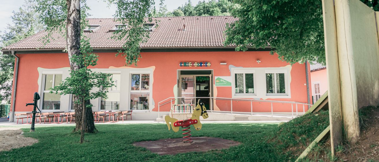 Ein orangefarbenes Gebäude mit weißen Rändern und einem braunen Dach steht vor einem Rasen mit einem gelben und roten Federpferdchen. Bäume und Tische mit Stühlen sind in der Nähe.