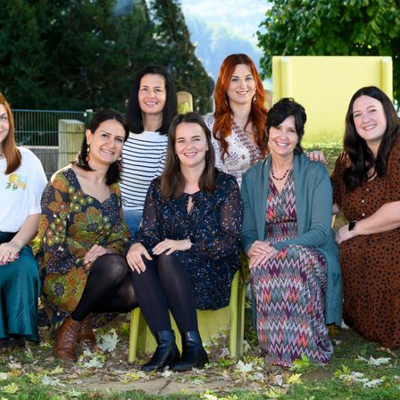 Sieben Frauen lächeln und posieren für ein Foto auf einer Wiese. Die Frau in der Mitte sitzt auf einem Stuhl. Hinter ihnen sind Bäume und ein Zaun.