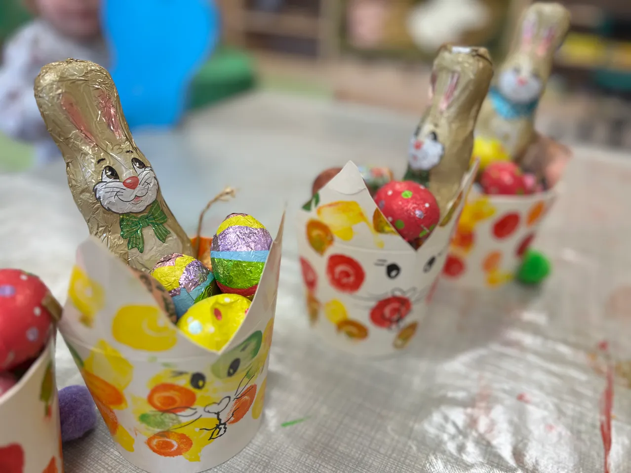 Drei Osterkörbchen mit jeweils einer goldenen Hasenfigur und verschiedenen Schokoladeneiern sind auf einem Tisch ausgestellt. Die Körbchen sind mit farbenfrohen Ostermotiven verziert.