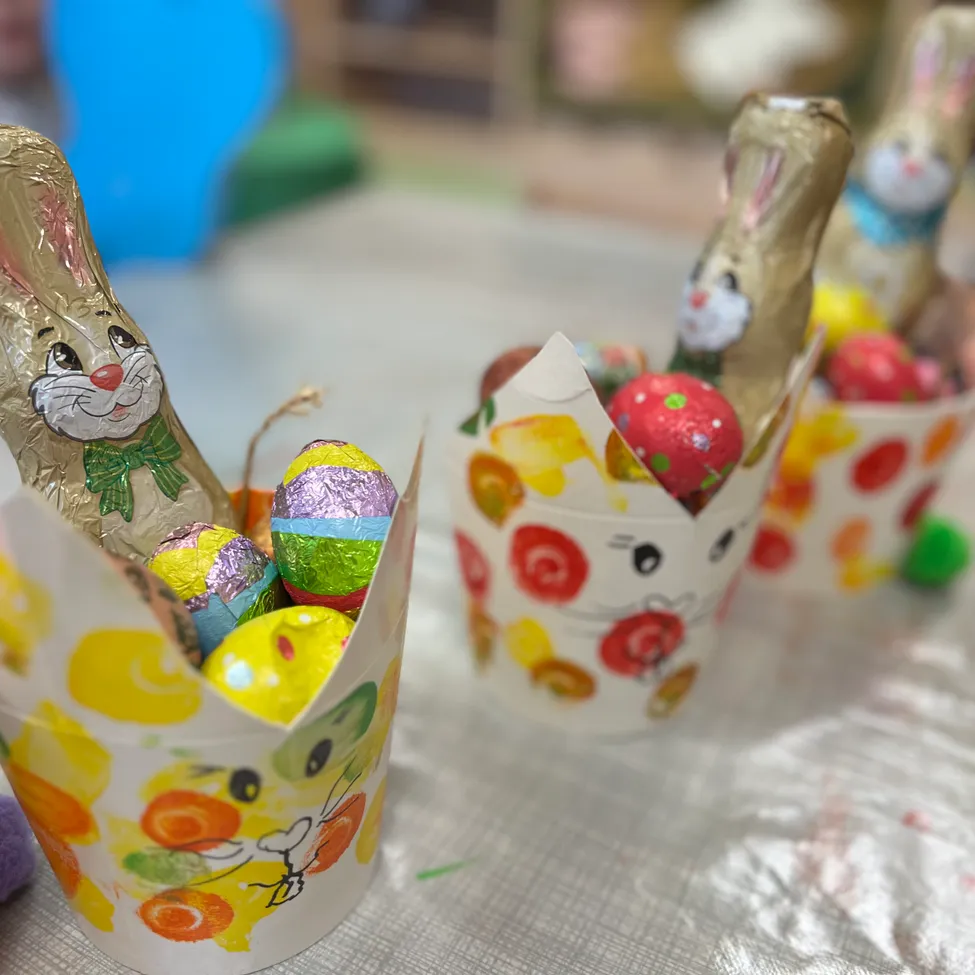 Drei Osterkörbchen mit jeweils einer goldenen Hasenfigur und verschiedenen Schokoladeneiern sind auf einem Tisch ausgestellt. Die Körbchen sind mit farbenfrohen Ostermotiven verziert.