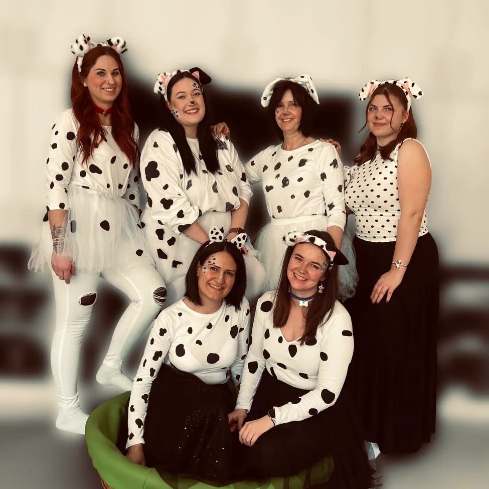 Eine Gruppe erwachsener Frauen in Dalmatiner-Kostümen, eine sitzt in einem grünen Korb, lächelt und posiert für ein Foto.