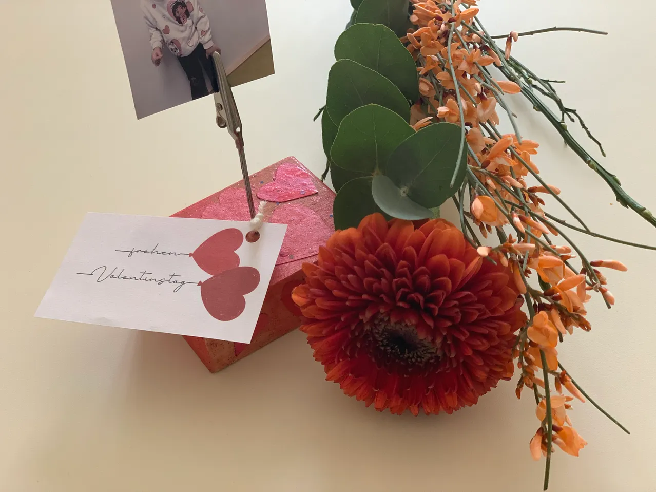 Ein Foto eines Kindes ist an einem roten Herzen auf einer rosa Box befestigt. Ein Valentinstagskarte mit Herzen ist an der Box befestigt. Eine orange Blume befindet sich neben der Box.