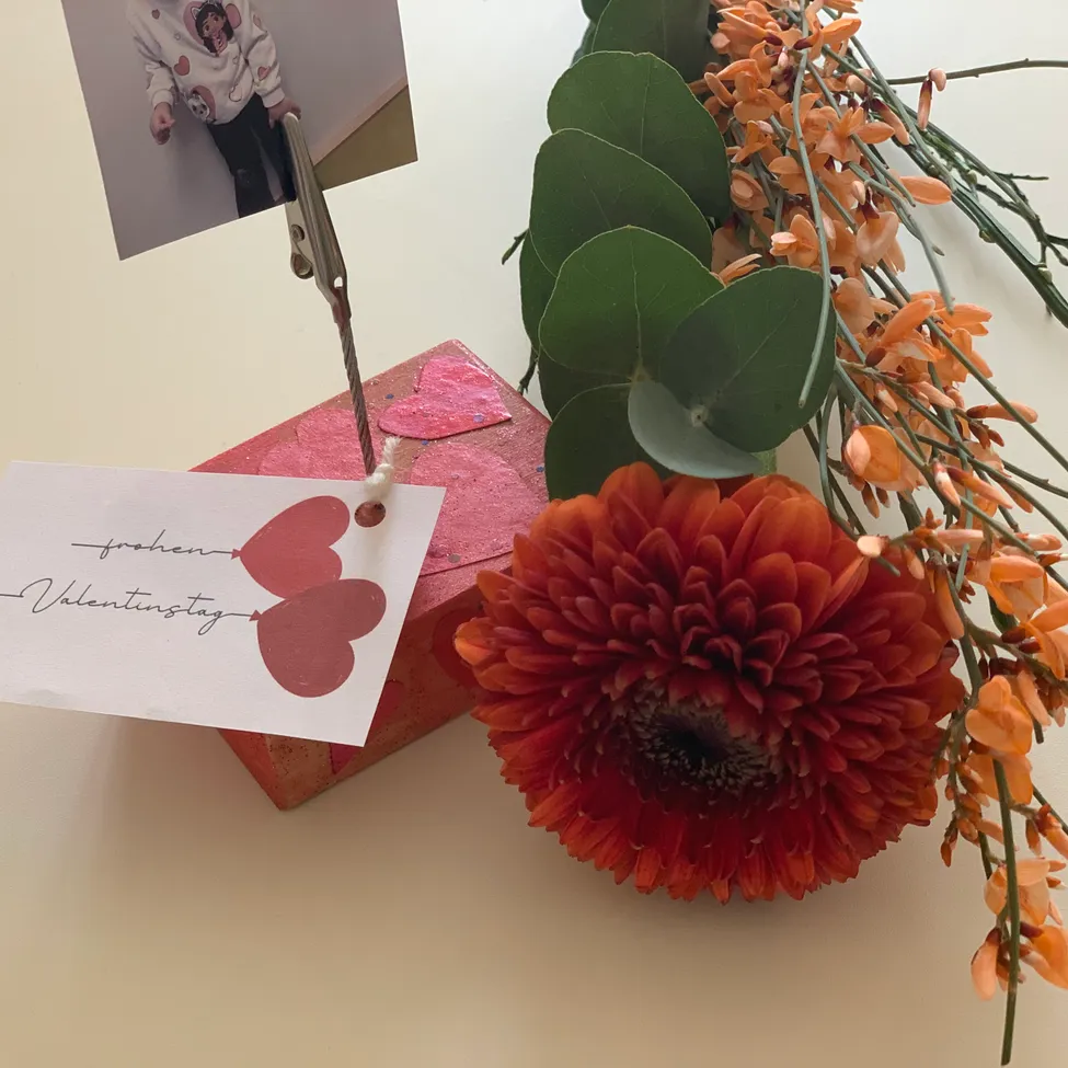 Ein Foto eines Kindes ist an einem roten Herzen auf einer rosa Box befestigt. Ein Valentinstagskarte mit Herzen ist an der Box befestigt. Eine orange Blume befindet sich neben der Box.