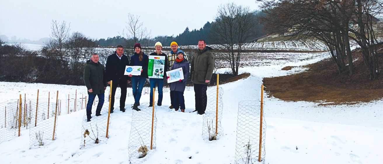Eine Gruppe von Menschen steht in einem verschneiten Feld und hält Schilder in der Hand. Hinter ihnen sind kahle Bäume und eine entfernte Landschaft zu sehen.