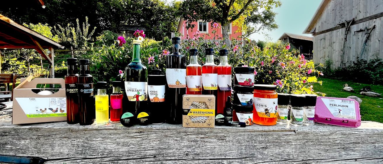 Ein Holztisch zeigt verschiedene Flaschen und Gläser, darunter Honig, Sirupe und andere Produkte, vor dem Hintergrund eines Gartens mit rosa Blumen und einem Baum.