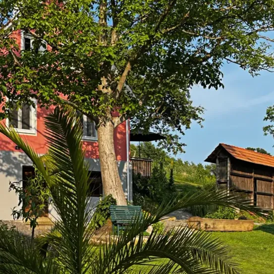 Ein rotes Haus mit weißen Fenstern befindet sich in einem üppigen grünen Garten mit einem Baum. Ein kleines Holzhaus steht in der Nähe, und eine grüne Bank befindet sich unter dem Baum.