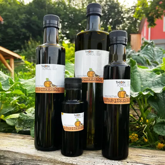 Vier Flaschen Kürbisöl sind auf einer Holzbank vor einem Hintergrund aus Grünzeug angeordnet. Jede Flasche hat ein Etikett mit dem Markennamen 'Thon Jorgl'.