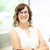 Eine Frau mit kurzen welligen braunen Haaren und einer Brille lächelt für ein Foto im Büro. Sie trägt ein weißes Oberteil, eine Halskette und eine Uhr. Sie verschränkt die Arme vor sich.