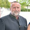 Drei Personen posieren im Freien mit einer Kulisse aus Bäumen und einem Gebäude. Eine Frau in einem blauen Kleid hält eine blaue Schriftrolle, während zwei Männer neben ihr lächelnd stehen.