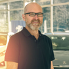 Ein Mann mit Brille und schwarzem Poloshirt steht lächelnd in einem Autohaus, mit Autos im Hintergrund.