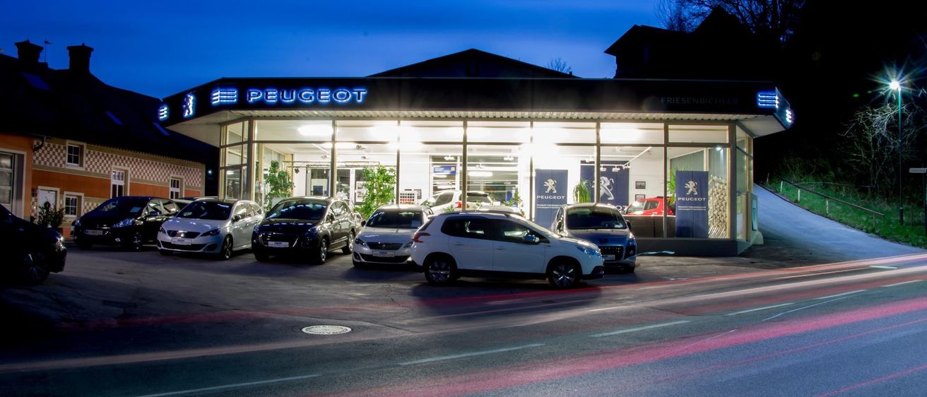 Ein Peugeot-Autohaus ist bei Nacht beleuchtet. Mehrere Autos sind davor geparkt. Das Schild über dem Eingang ist blau mit weißen Buchstaben.