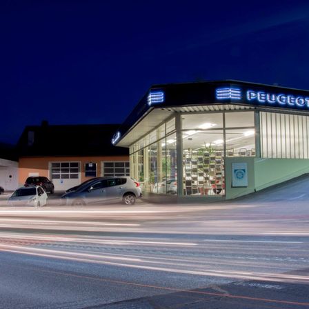 Peugeot-Autohaus bei Nacht mit geparkten Autos davor und Lichtern im Inneren. Fahrzeuge in Bewegung verschwimmen im Vordergrund.