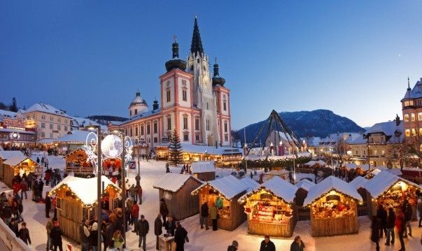 Outdoor-Weihnachtsmarkt mit Holzhütten auf einem verschneiten Platz, Stadtkirche im Hintergrund. Leute erkunden Stände, beleuchtet von warmen Lichtern.