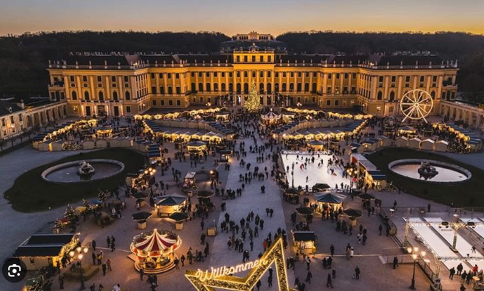 Luftaufnahme eines Weihnachtsmarktes vor einem großen Palast mit vielen Menschen, einem Riesenrad und einer Eislaufbahn.