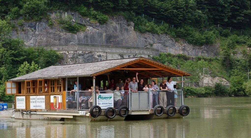 Eine Gruppe von Menschen auf einem überdachten Boot, das auf einem Fluss mit einer felsigen Klippe im Hintergrund fährt.