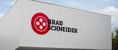 Die Außenseite eines großen weißen Gebäudes mit einem roten Logo und dem Wort Brau Schneider. Es ist von Kies und einem Schild umgeben. Bäume und ein Auto sind im Hintergrund zu sehen.