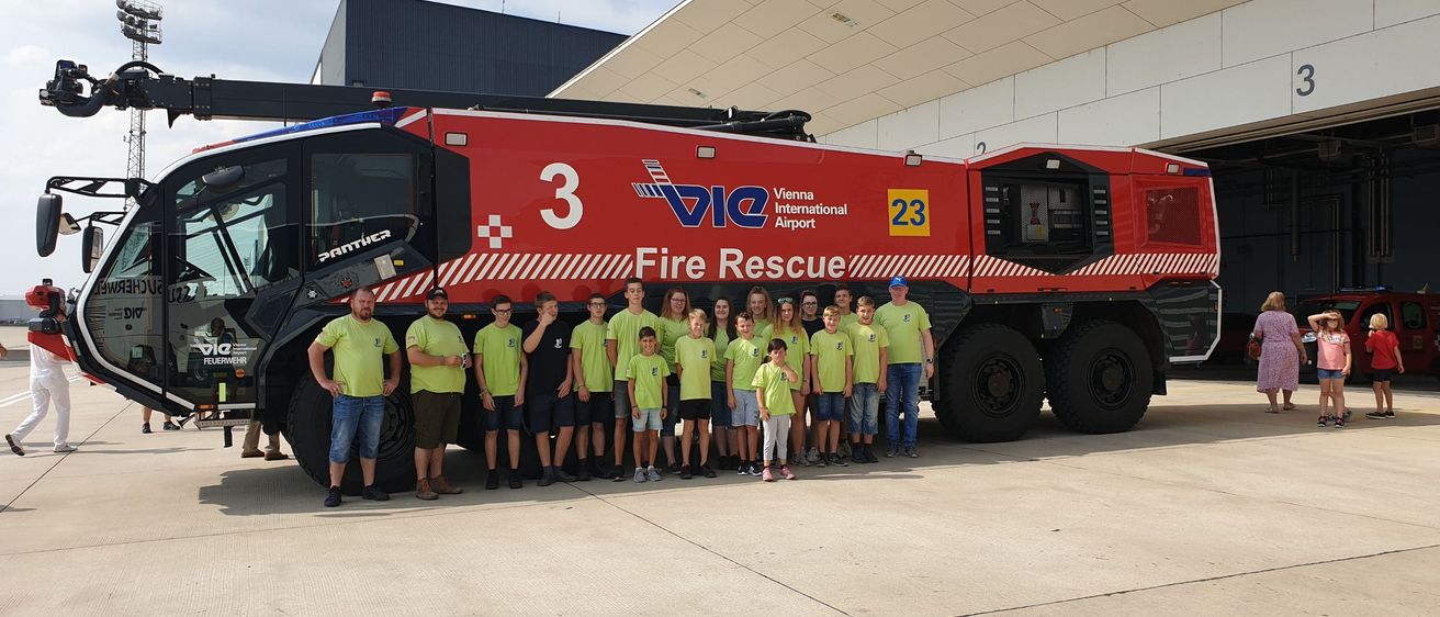 Eine Gruppe von Kindern und Erwachsenen in grünen Shirts steht vor einem roten Feuerwehrauto mit der Aufschrift VIC 3. Das Auto ist vor einem Gebäude geparkt.