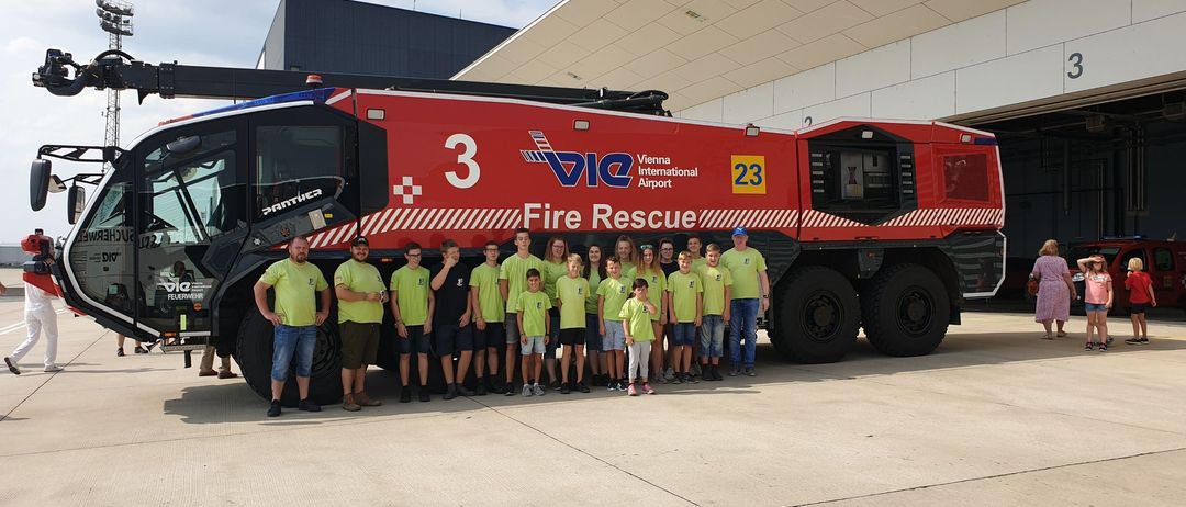 Eine Gruppe von Kindern und Erwachsenen steht vor einem großen roten Feuerwehrauto, das mit Vienna International Airport und Nummer 3 gekennzeichnet ist.