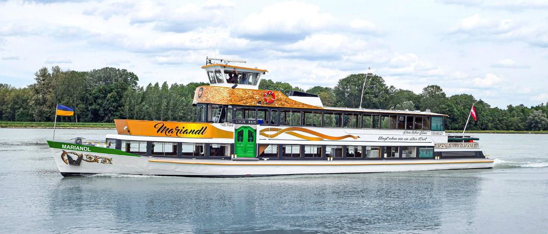 Eine Fähre namens 'Mariandl' fährt auf einem Fluss mit Bäumen im Hintergrund. Die Fähre hat ein goldenes Dach und grüne Türen. Sie kann 148 Personen befördern.