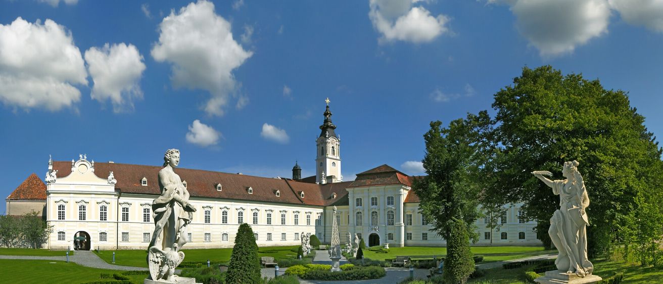 Ein großes Barockschloss mit Statuen im Garten unter teilweise bewölktem Himmel.