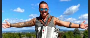 A man stands with an accordion, arms wide open, smiling in a sunny outdoor area with trees and houses in the background.