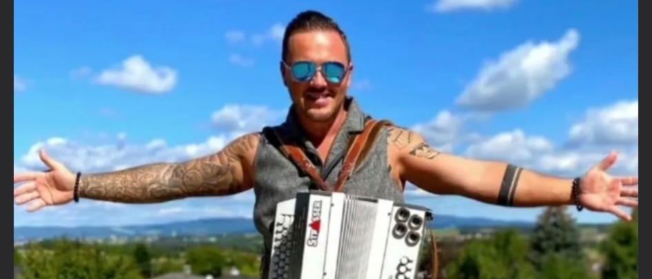 Ein Mann steht mit einem Akkordeon, Arme weit geöffnet, lächelnd in einem sonnigen Außenbereich mit Bäumen und Häusern im Hintergrund.