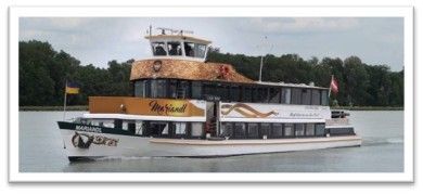 Ein großes weißes und orangefarbenes Boot, mit der Aufschrift 'Mermaid', fährt auf einem Fluss mit Bäumen im Hintergrund. Eine Flagge ist am Bug des Bootes sichtbar.