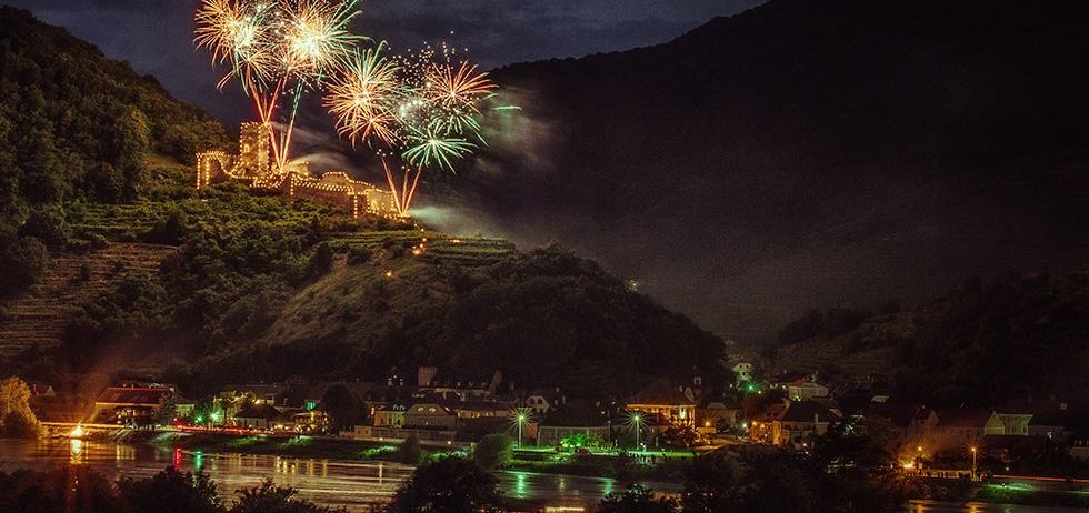 Ein Nachtblick auf eine Stadt mit Feuerwerk über einem Berg und einem Fluss, beleuchtet von Lichtern.