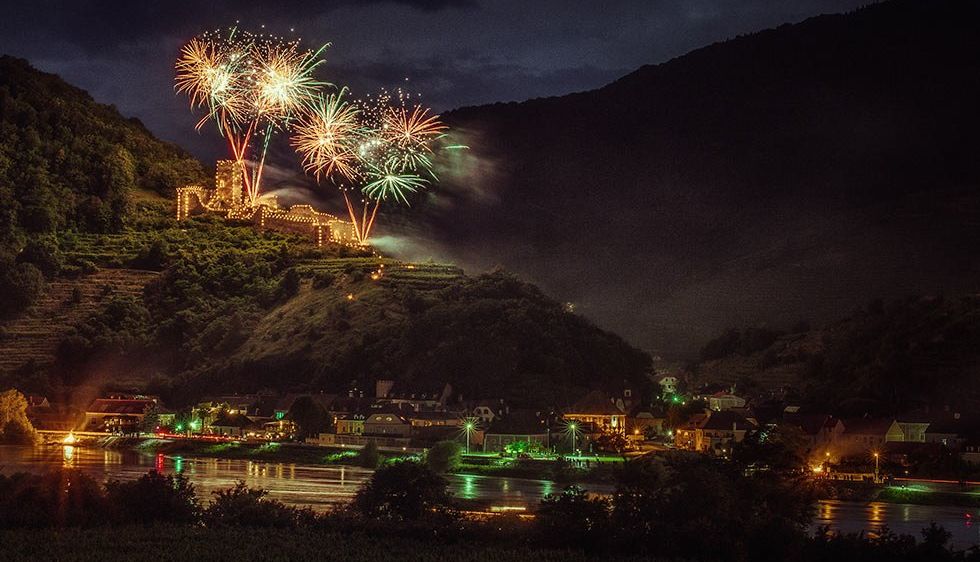 Ein Nachtblick auf eine Stadt mit Feuerwerk über einem Berg und einem Fluss, beleuchtet von Lichtern.