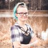 A woman with blue hair and glasses stands near a lake. She wears a checkered shirt and has a tattoo on her arm.