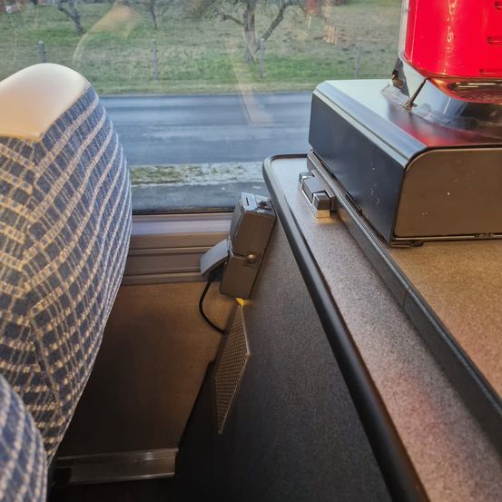 Im Inneren eines Zuges zeigt ein Fenster eine Straße mit einem grasbewachsenen Feld. Eine rote Notleuchte befindet sich auf einem grauen Regal. Ein blau gemusterter Sitz und ein grauer Armlehnen sind zu sehen.