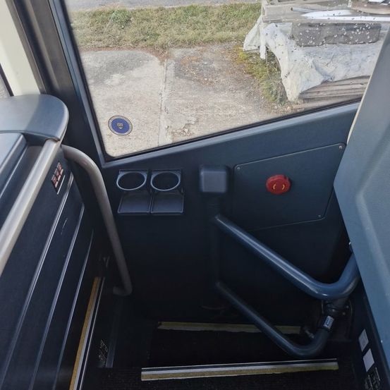 Der Blick von innen in einem Bus zeigt den Fahrerbereich mit einem roten Knopf, Getränkehaltern und einem kleinen Fenster mit Logo. Die Außenansicht ist von einem Betonbereich mit Gras.